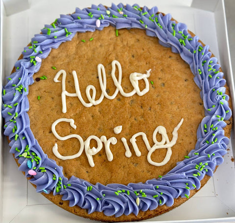 Close-up of a custom gluten-free cookie cake with lavender buttercream frosting, green sprinkles, and a hand-piped “Hello Spring” message.