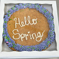 Round gluten-free cookie cake with purple buttercream frosting, green sprinkles, and a piped “Hello Spring” message, packaged in a bakery box from Sinful Confections.